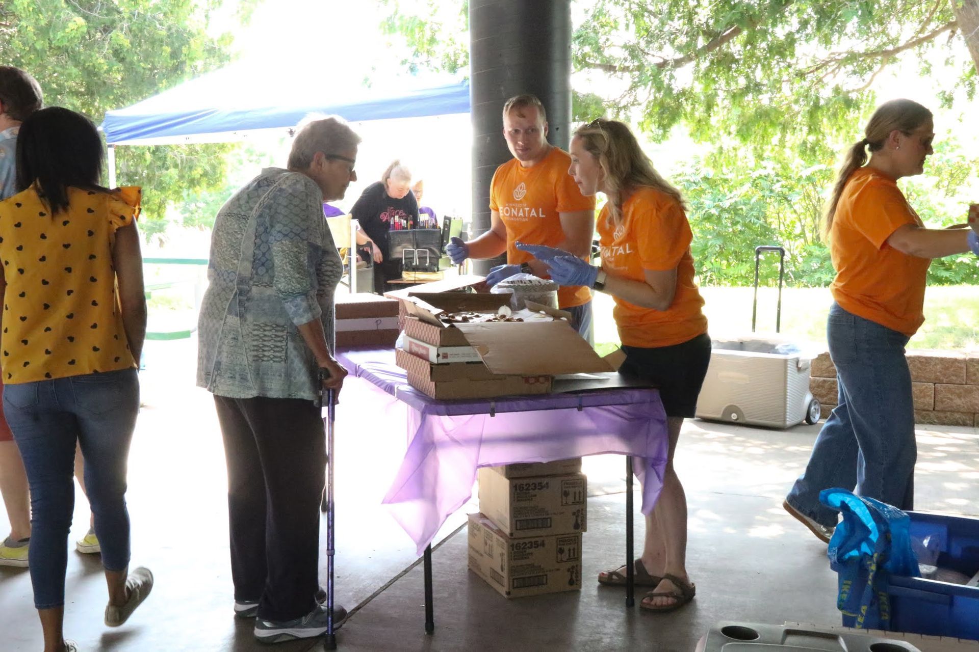 People volunteer at a table with food under a pavilion, handing items to others.