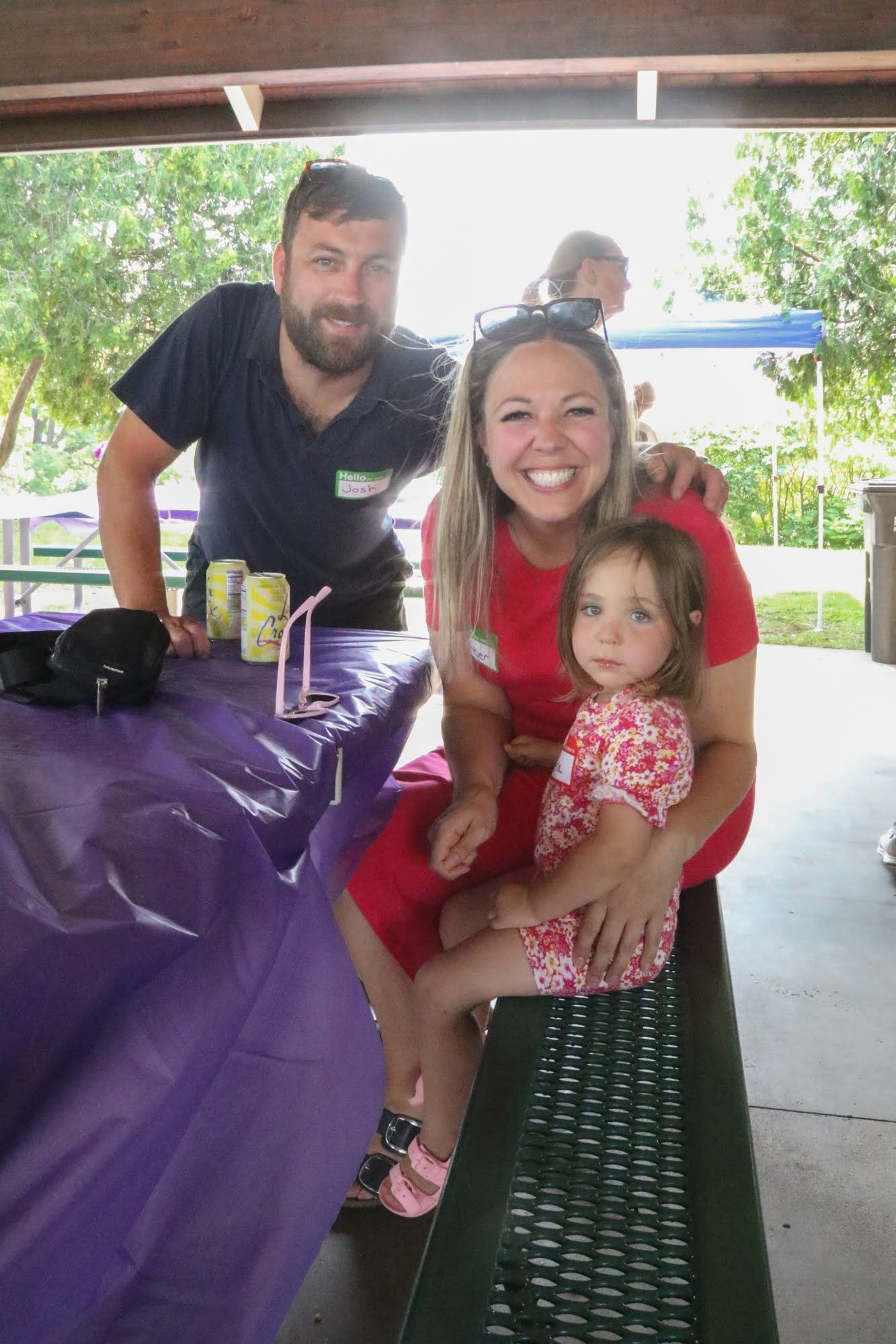 Family of three at a picnic table: adult woman holding a child smiles; man stands behind table.