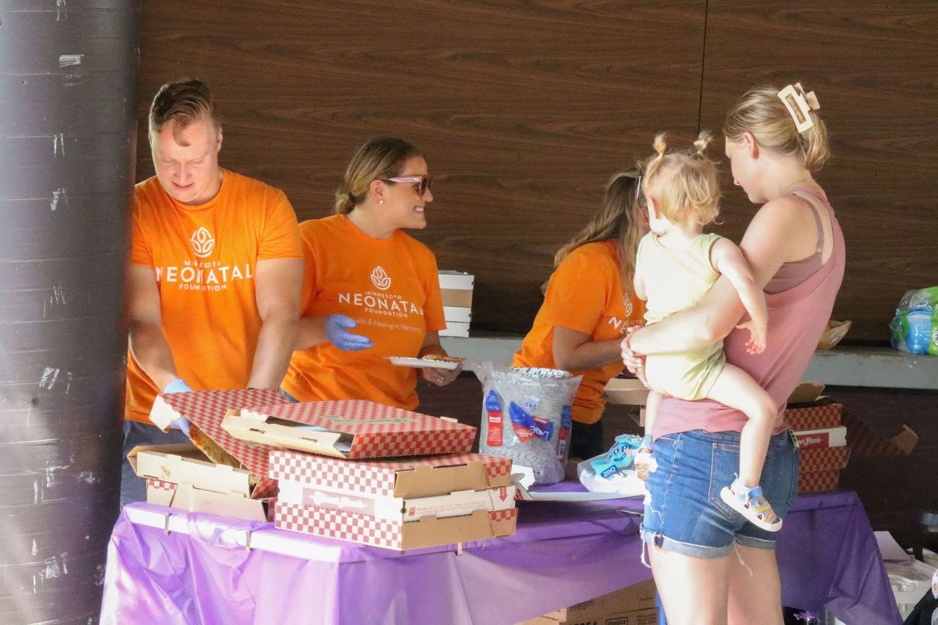 People serving food at an outdoor event. Servers wear orange shirts. A woman holds a toddler.