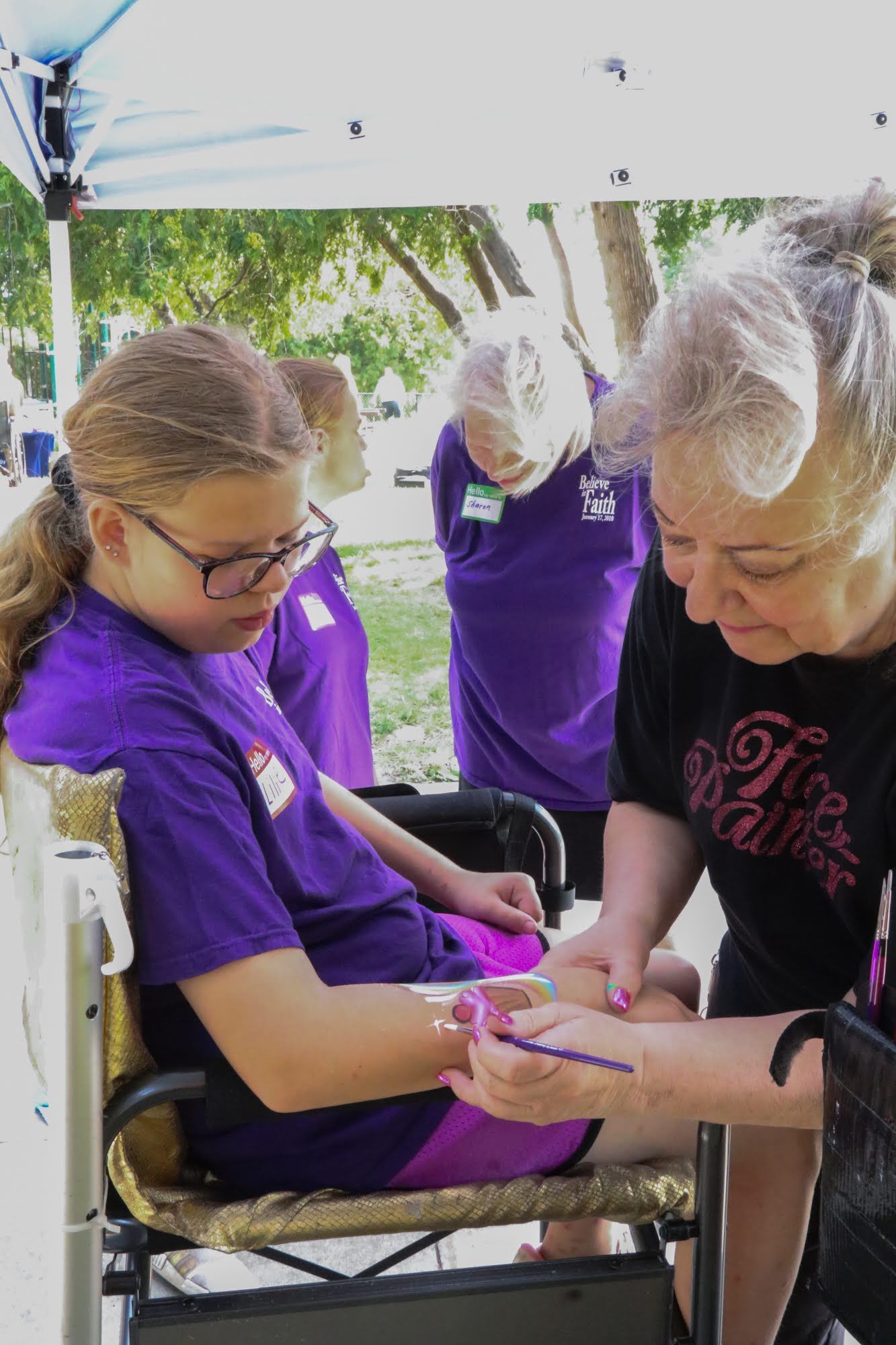 Person in wheelchair getting face painting at an outdoor event. Another person paints her arm. People in purple shirts watch.