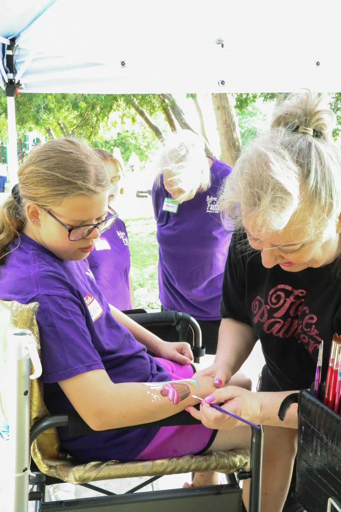 Girl in wheelchair getting face painting at an outdoor event, with two women watching.
