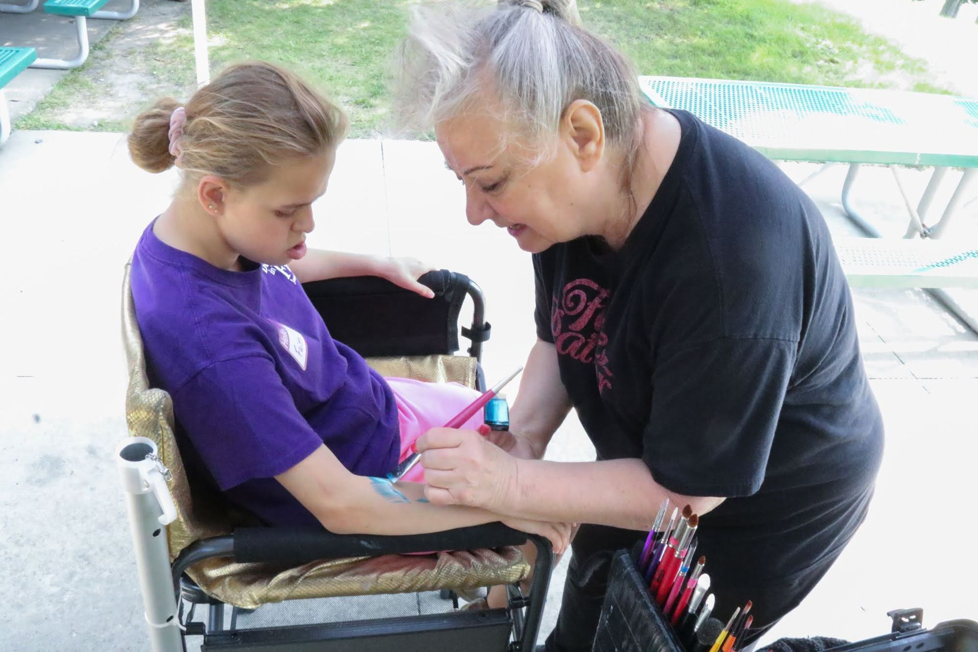 Woman paints child's arm while child sits in wheelchair outdoors.