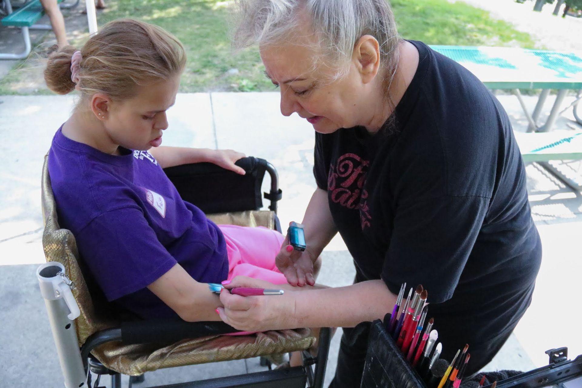 Woman painting a design on a person's arm, who is seated in a wheelchair outdoors.