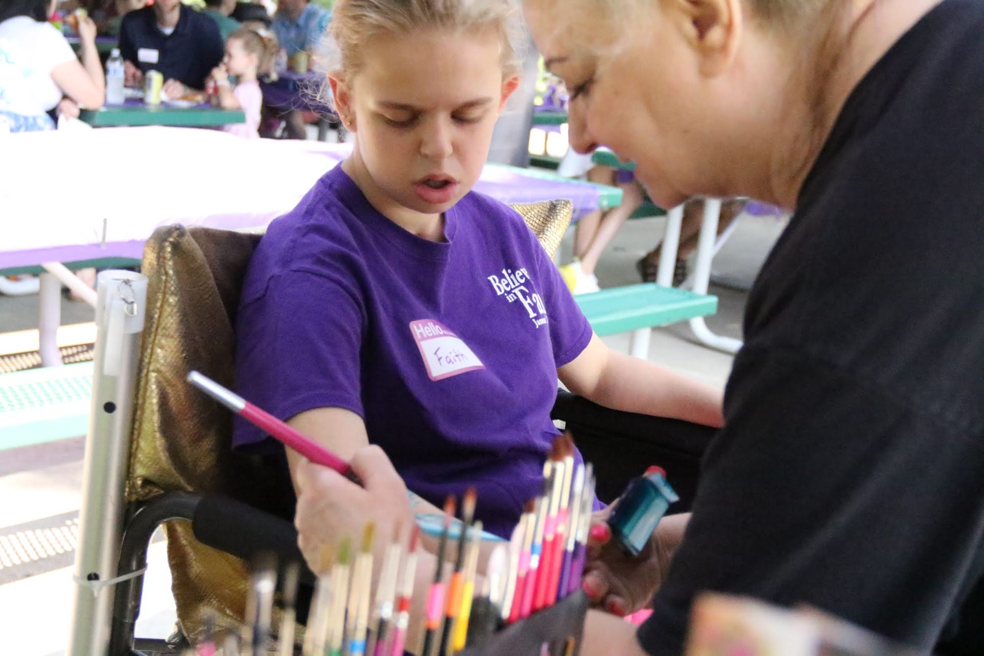 Woman in wheelchair being assisted with a paintbrush, seated at a table with other people.