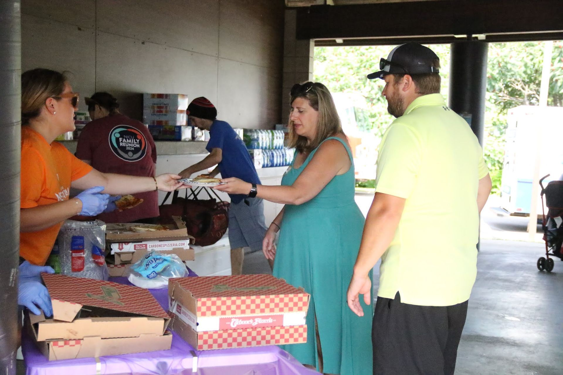 People receiving food from volunteers at an outdoor event. Pizza boxes, water bottles, and a stroller are visible.