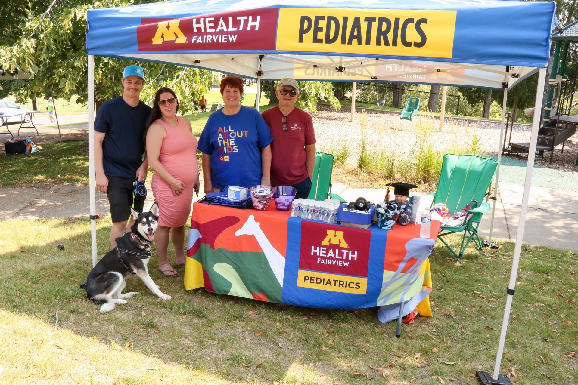 People at a booth for M Health Fairview Pediatrics. Colorful banner, dog, sunny outdoor setting.