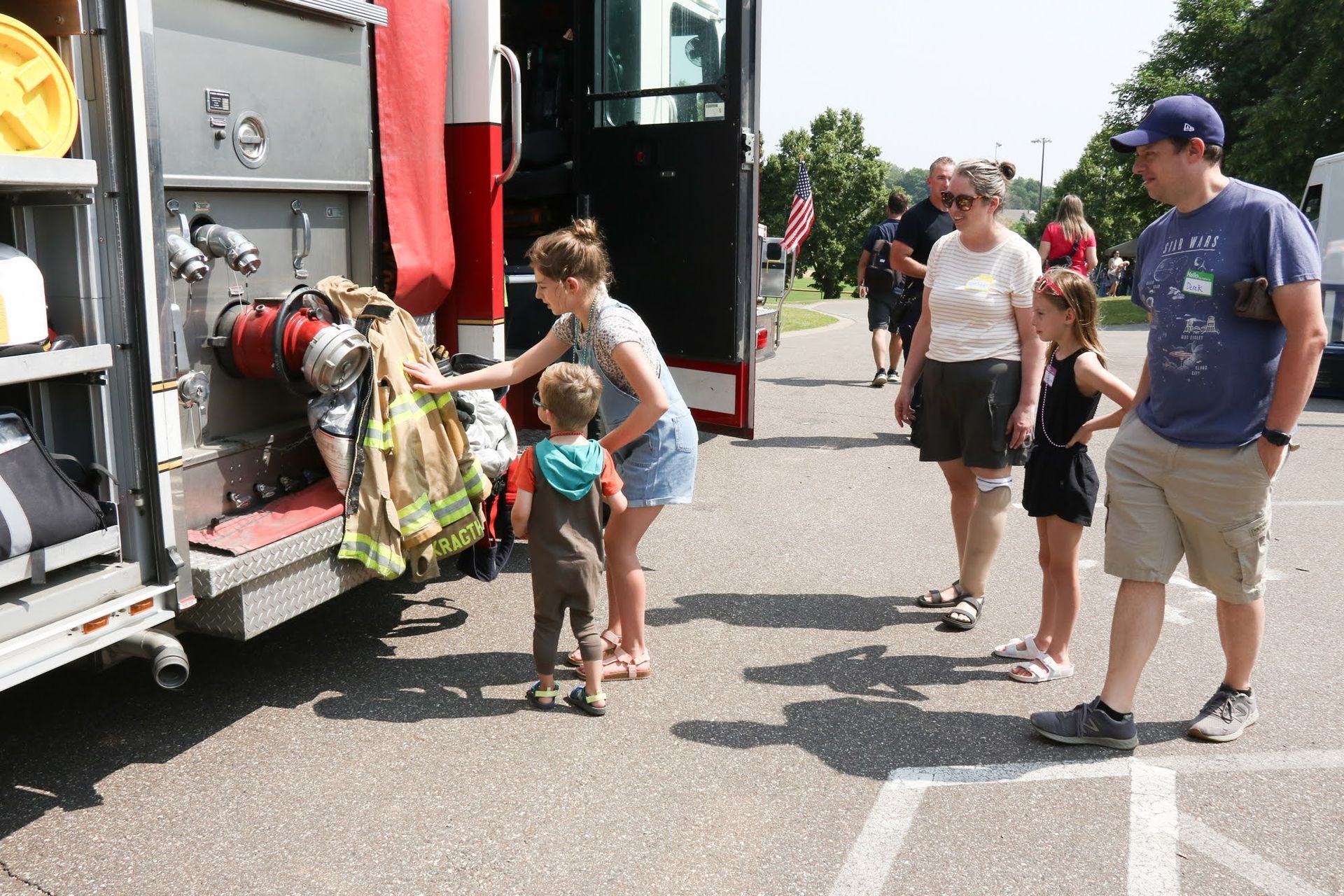 Family exploring a fire truck. A woman helps a child with gear. Others watch nearby. Outdoors in sunlight.