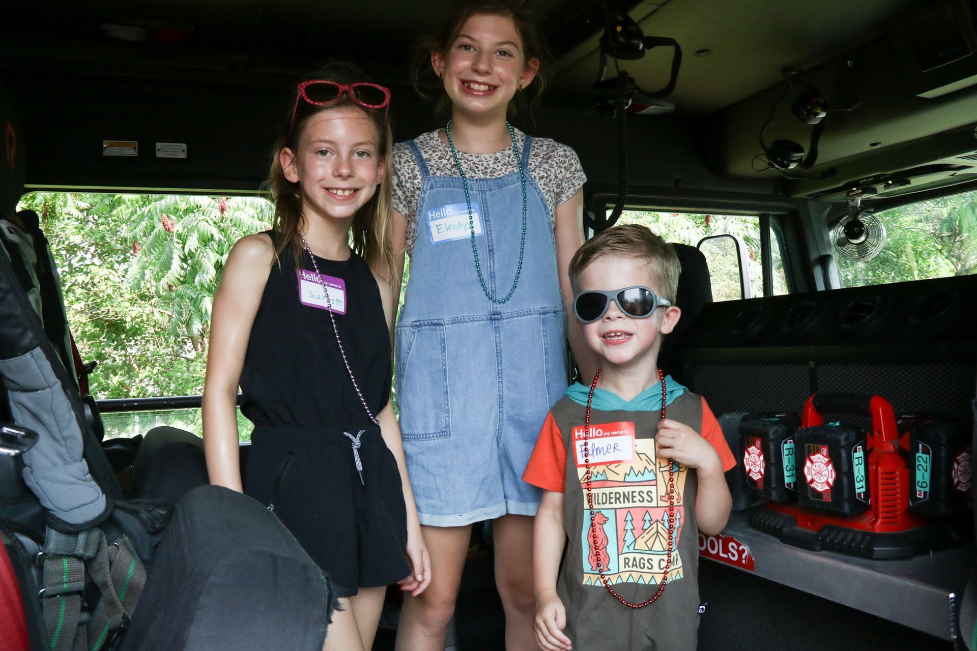 Three children inside a vehicle, smiling. Two girls and a boy wear sunglasses and name tags.
