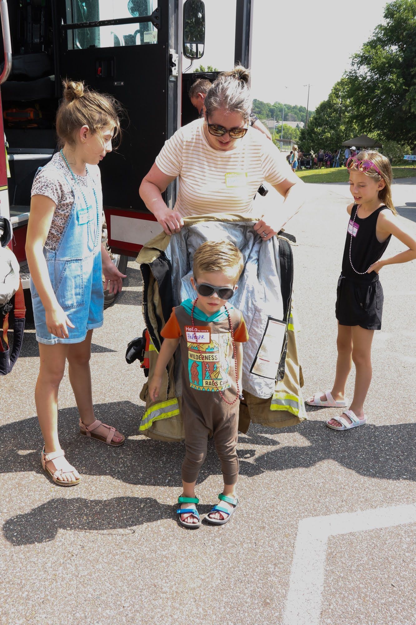 A child in sunglasses tries on fire gear, assisted by an adult and observed by two children near a fire truck.