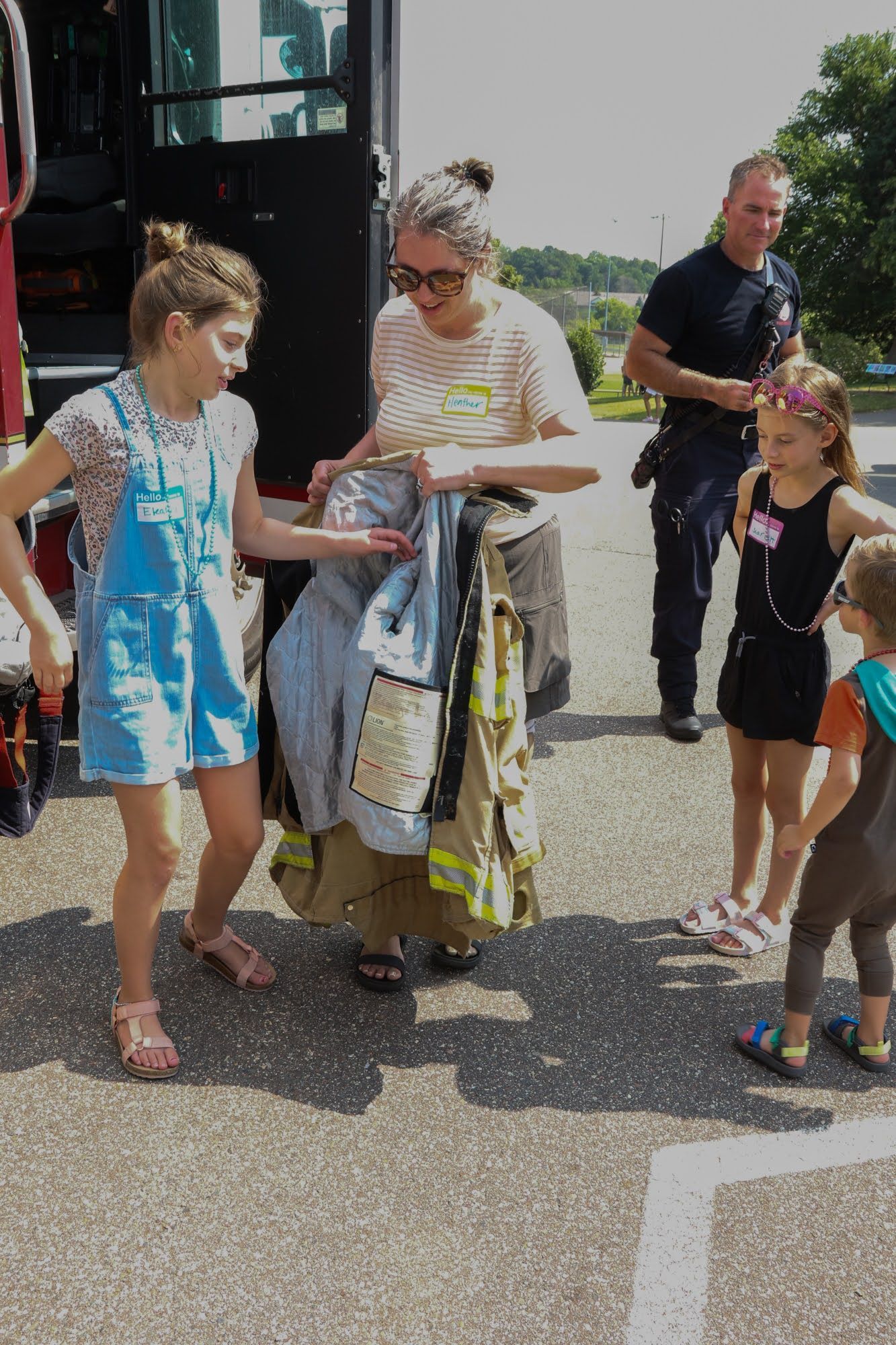 Woman holding firefighter gear, showing children near fire truck. Another firefighter watches.