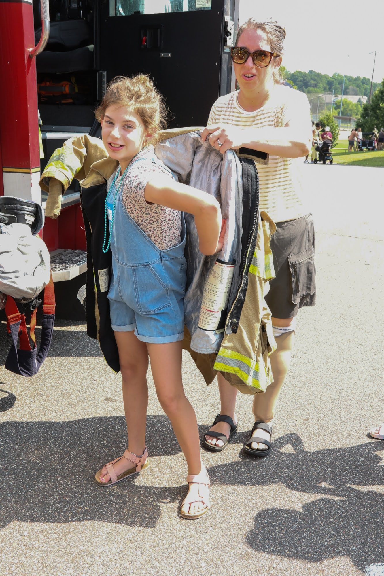Girl in overalls trying on firefighter gear with a woman nearby, standing next to a fire truck.