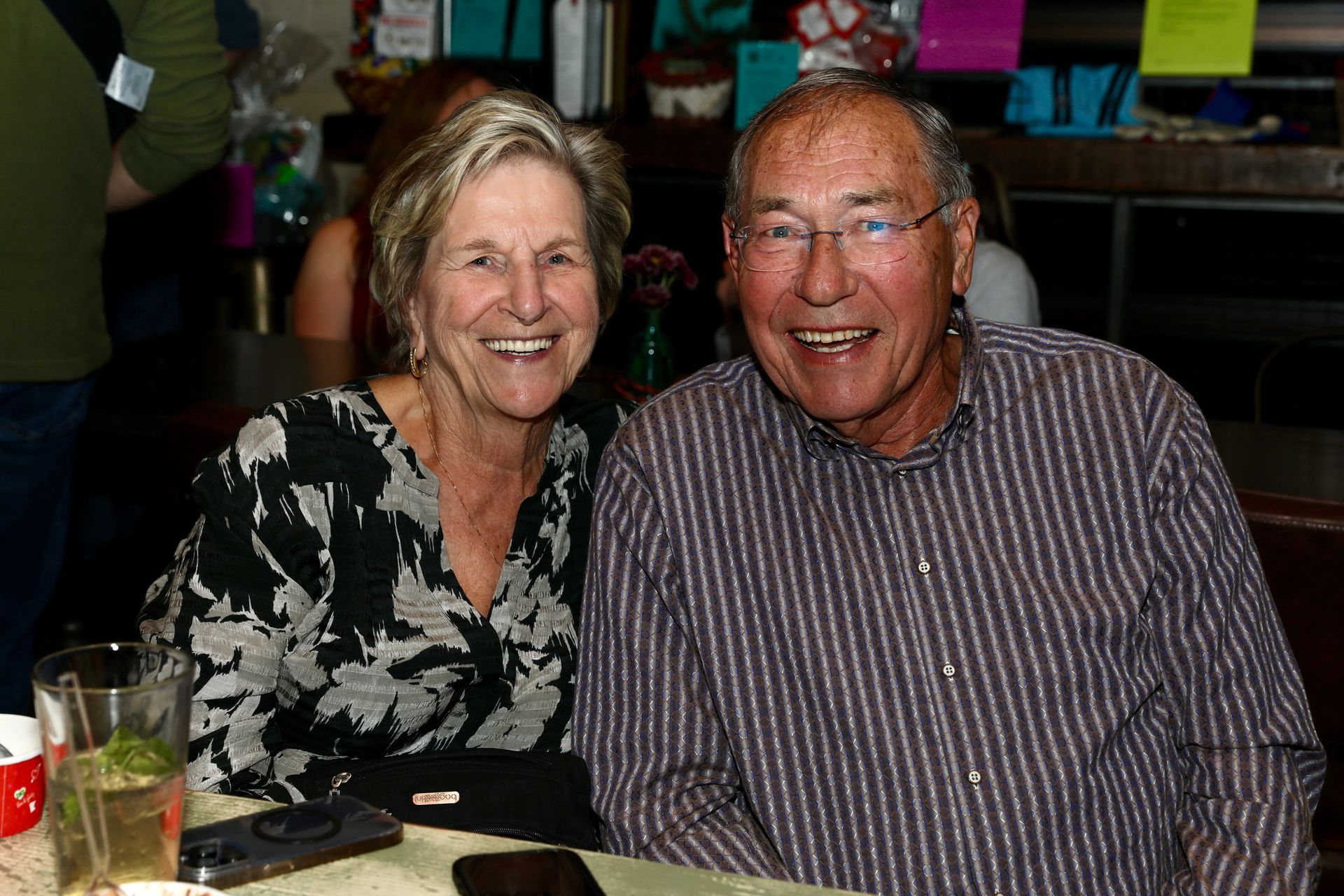Smiling older couple sitting at a table. Woman in floral blouse, man in striped shirt.