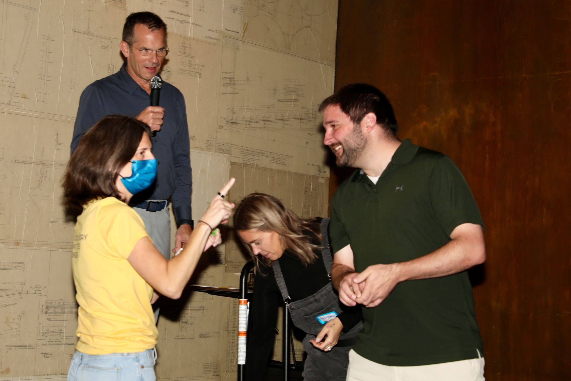Four people on stage; one with a microphone. Woman in yellow shirt points up at man. Other man laughs. Dark background.