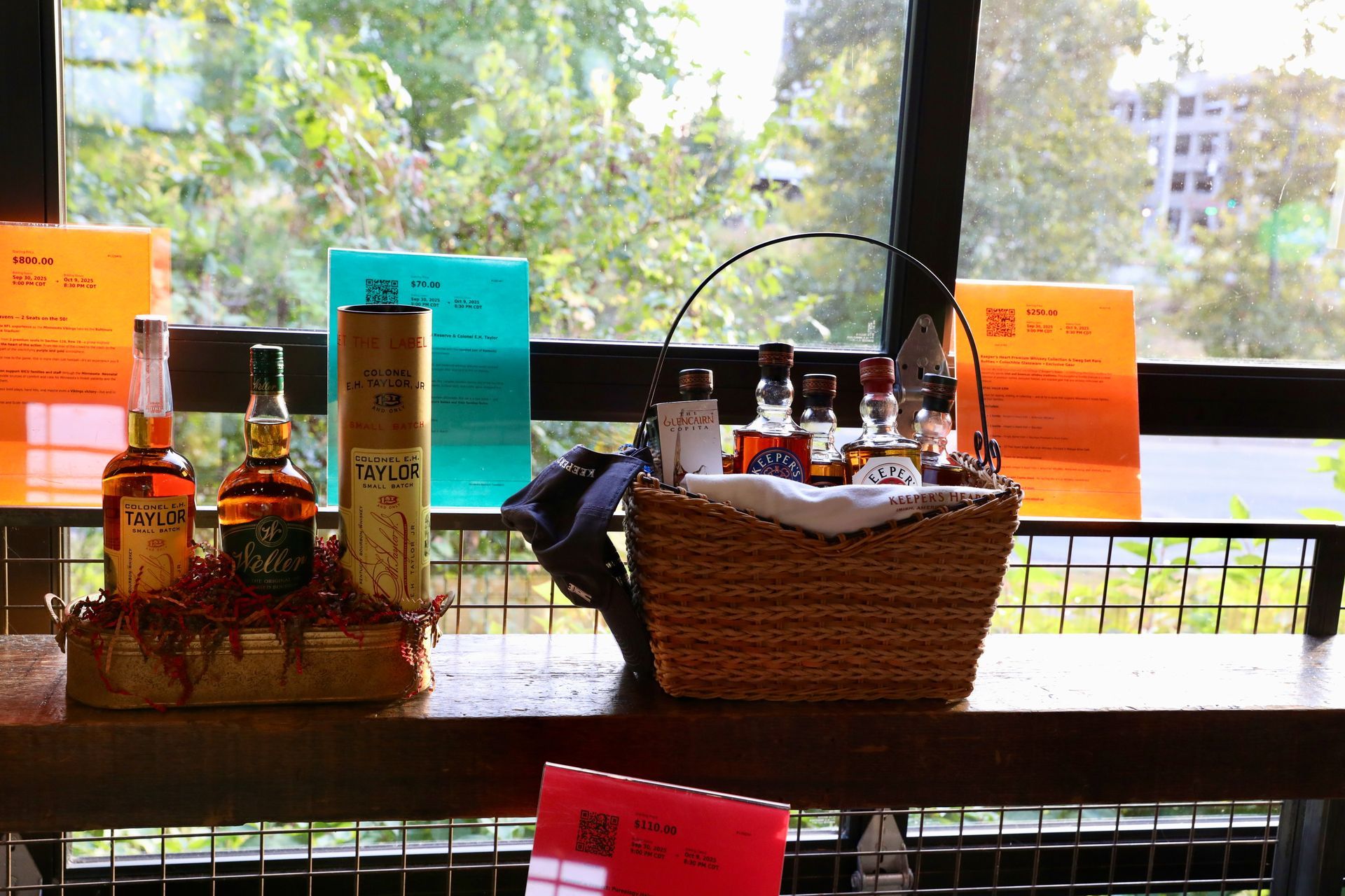 Whiskey bottles and a gift basket on a table, with a view of trees through a window.