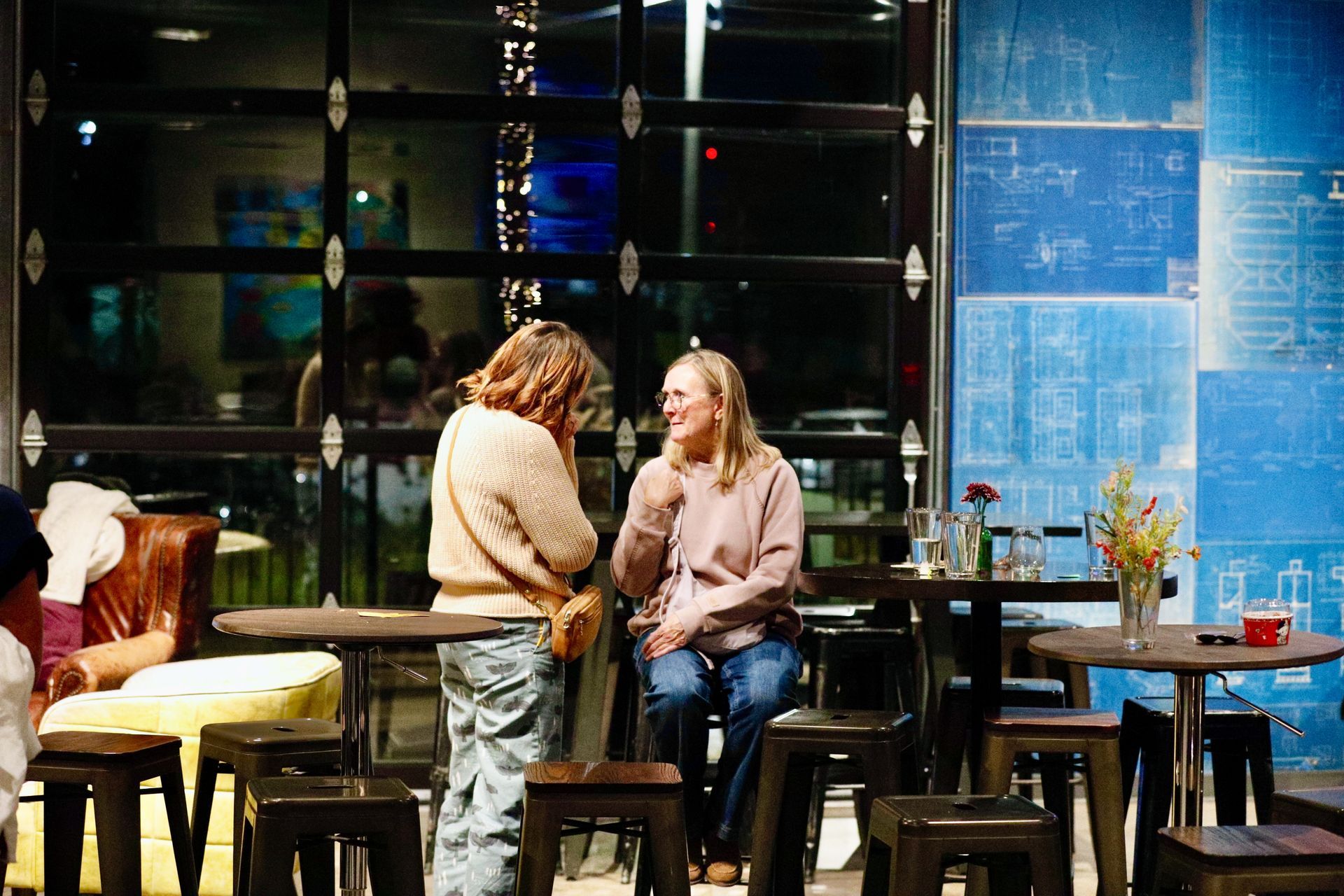 Two people conversing in a cafe. One standing, other seated. Blue blueprints on the wall, dark interior with outdoor view.