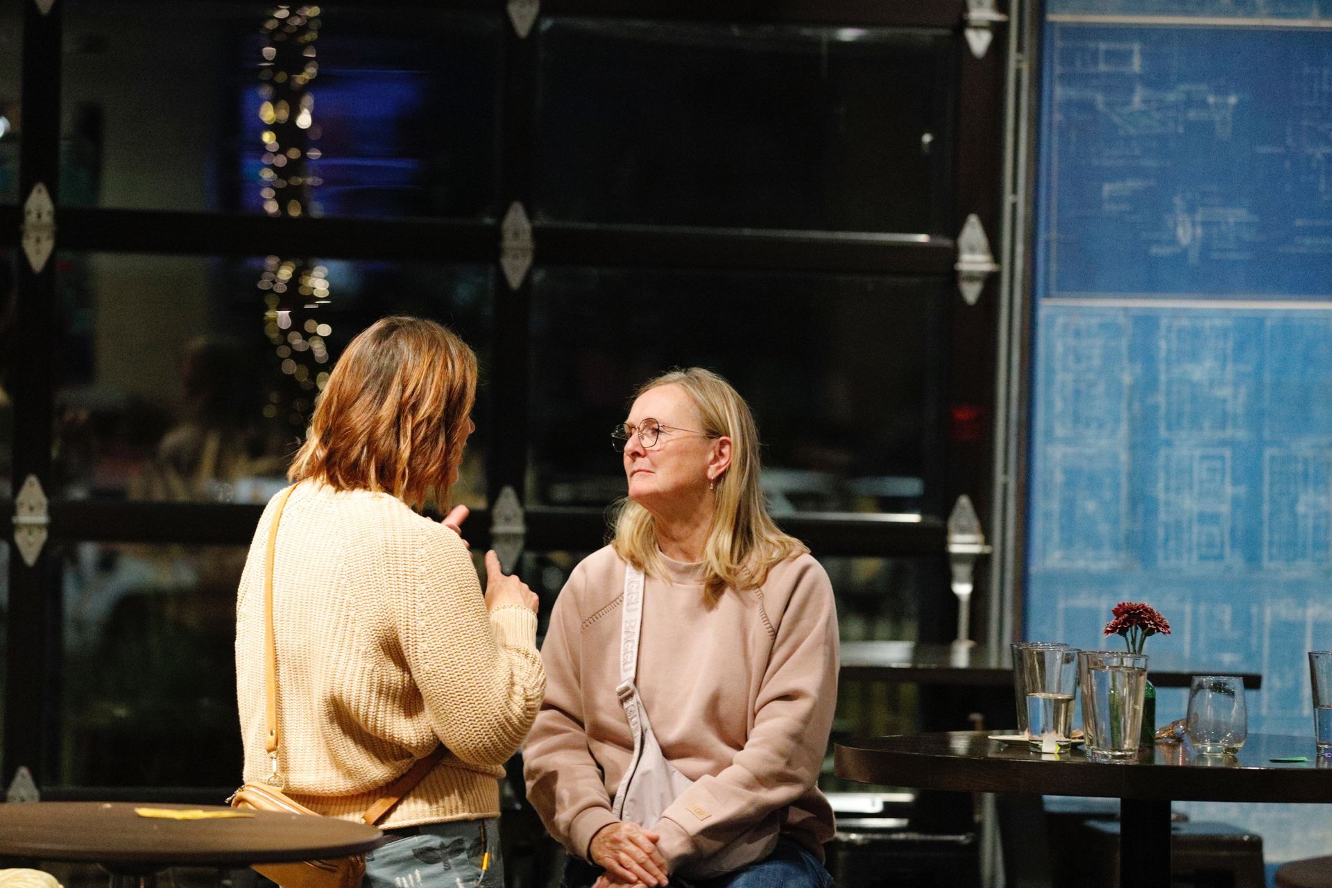 Two women talking in a dimly lit room, one holding a microphone. Blue blueprint-style art and a glass-paned door are visible.