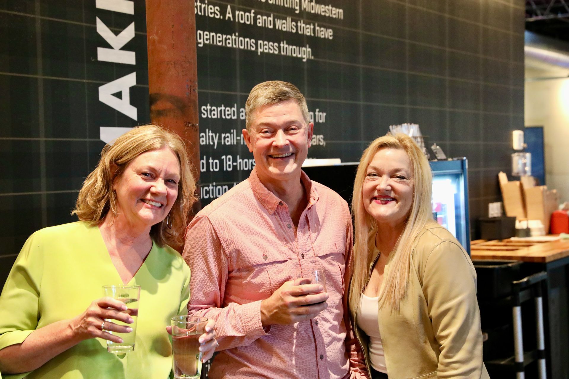 Three people smiling, holding drinks, in front of a wall with text; a man in pink shirt is flanked by women in yellow and tan.