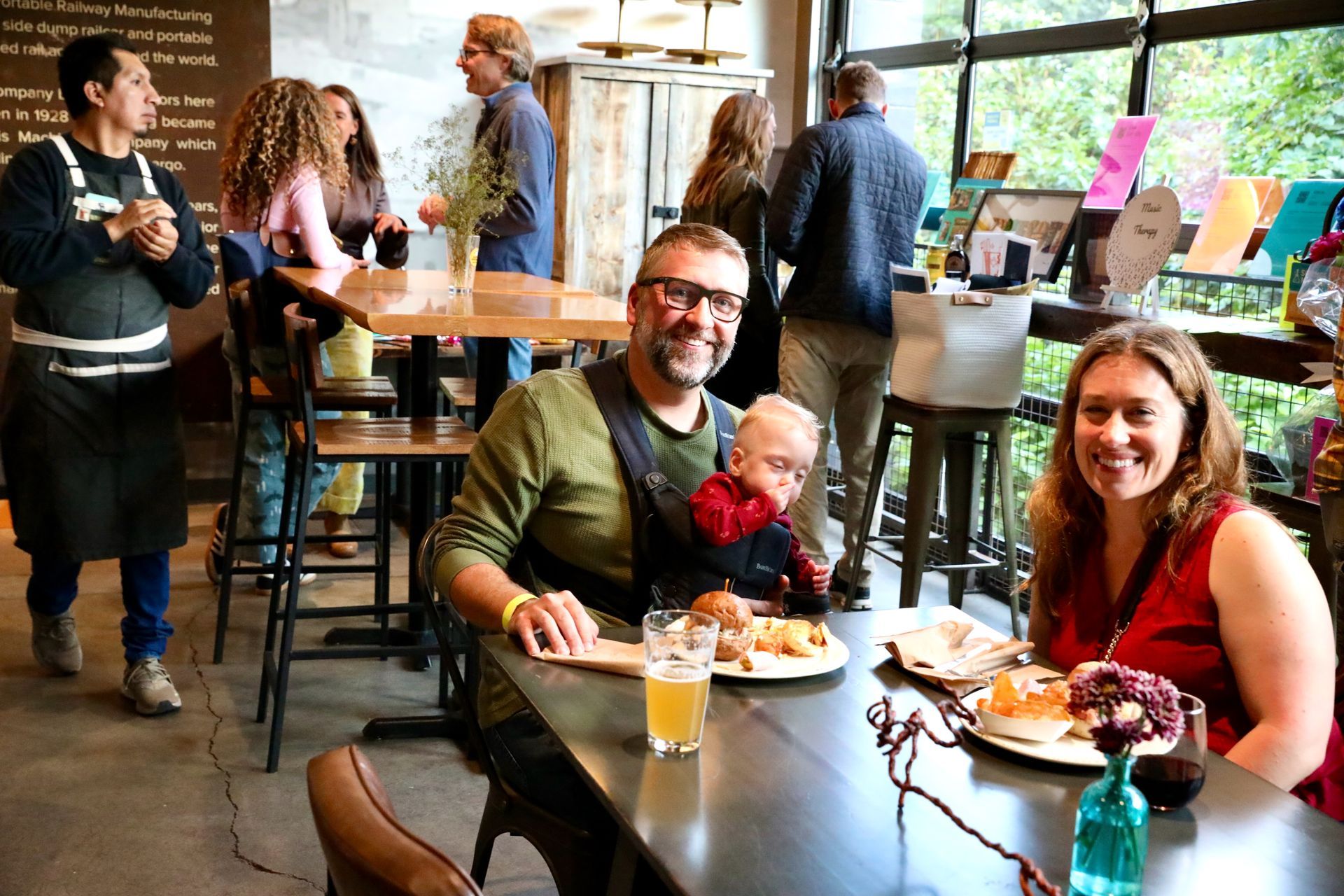 Family at a brewery: man with baby in carrier, woman, food, drinks on table, other patrons in background.