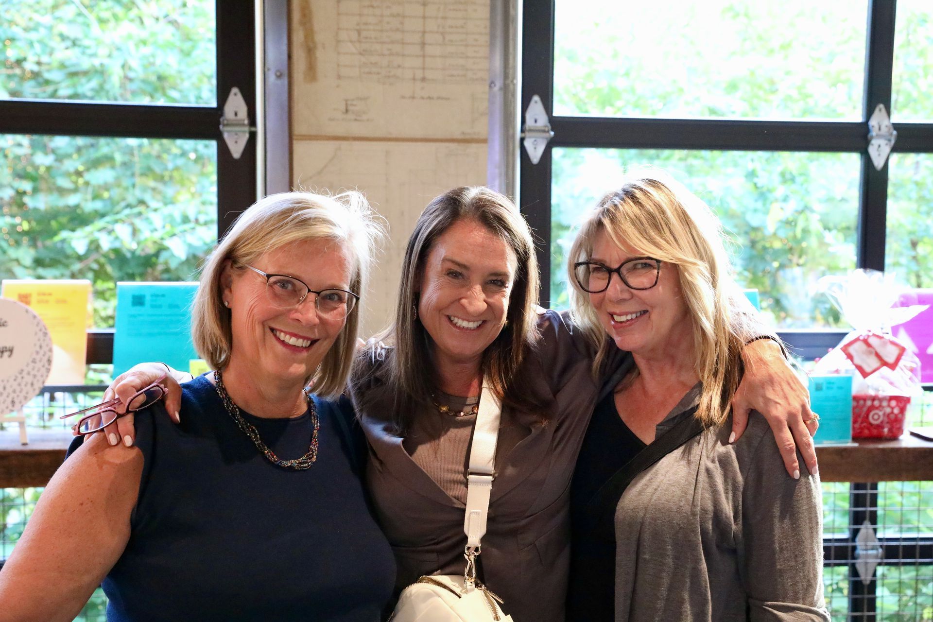 Three smiling women with arms around each other pose indoors near a window.