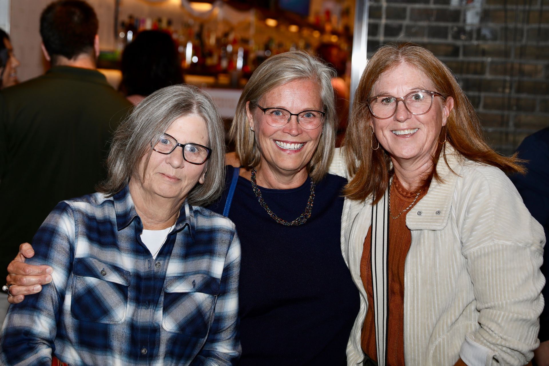 Three people smiling, posed together indoors. One in plaid shirt, one in a blue top, and one in a cream-colored jacket.