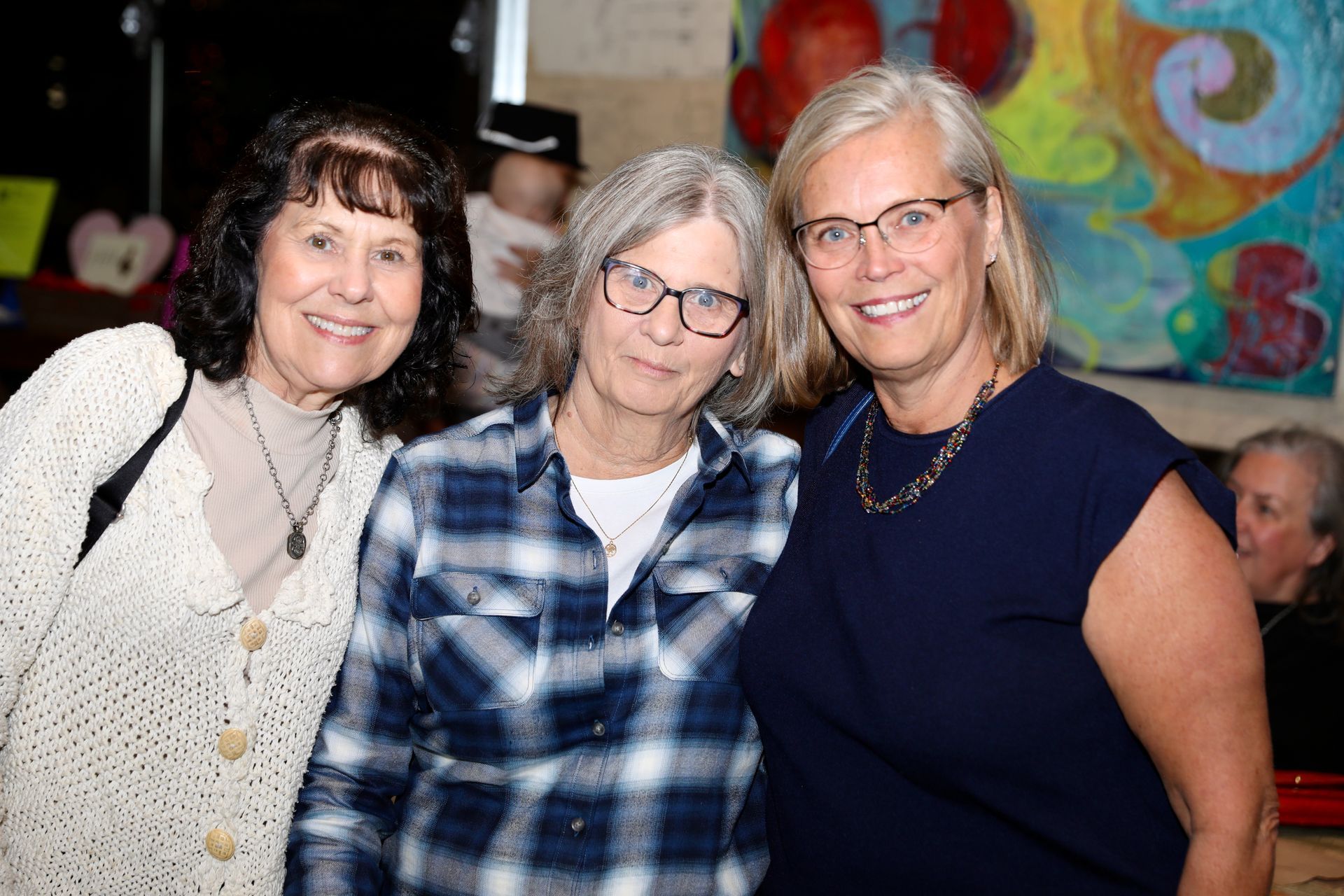 Three women smiling, posing for a photo indoors, one in blue, one in plaid, one in beige cardigan.