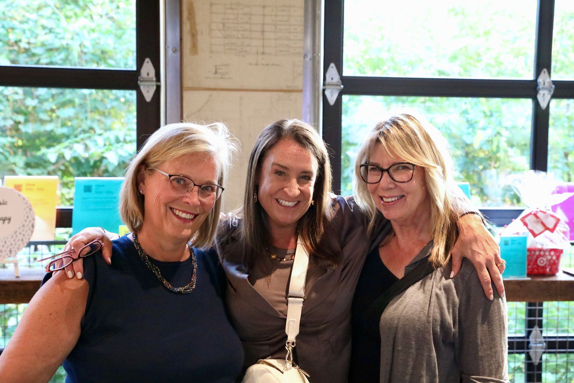 Three women smiling, arms around each other. Inside, near a window, with items on a table.
