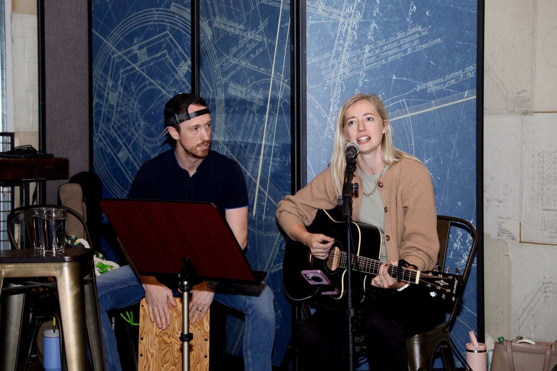 Two musicians performing: woman singing and playing guitar, man with percussion instrument. Blue blueprint background.