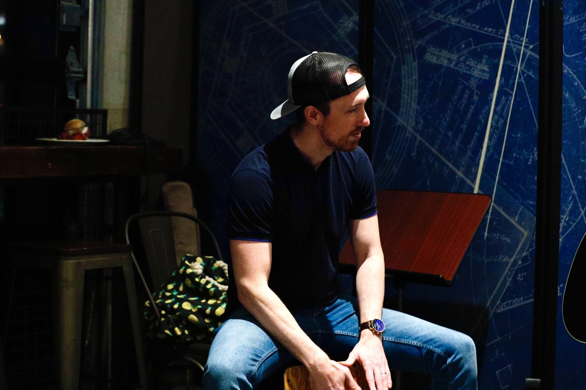 Man in a cap and blue shirt sits, looking to the side, in front of a blue wall with geometric patterns.