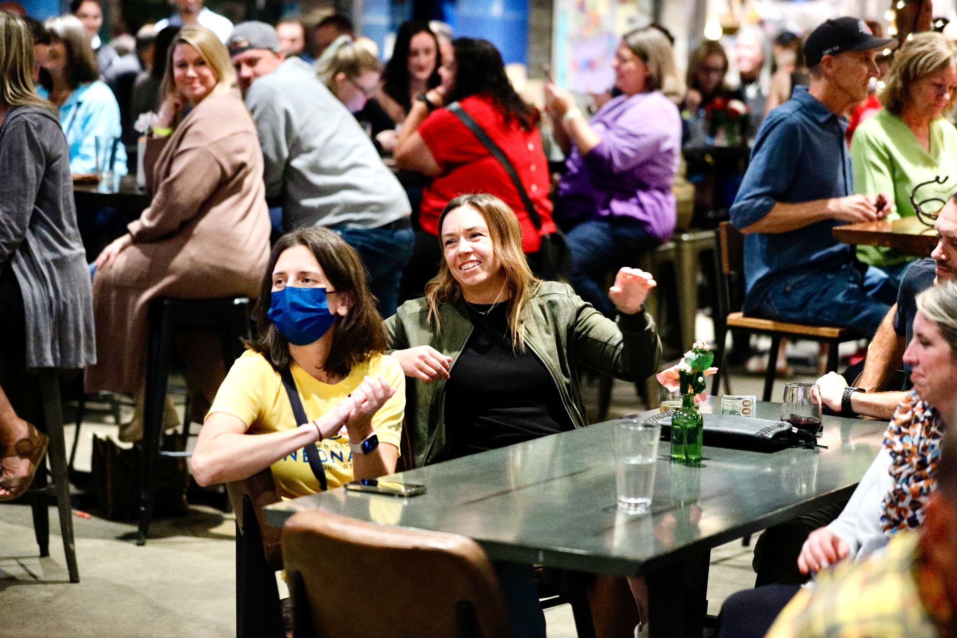 People at a table inside, some smiling, one wearing a mask, others in background, sitting at tables, listening.