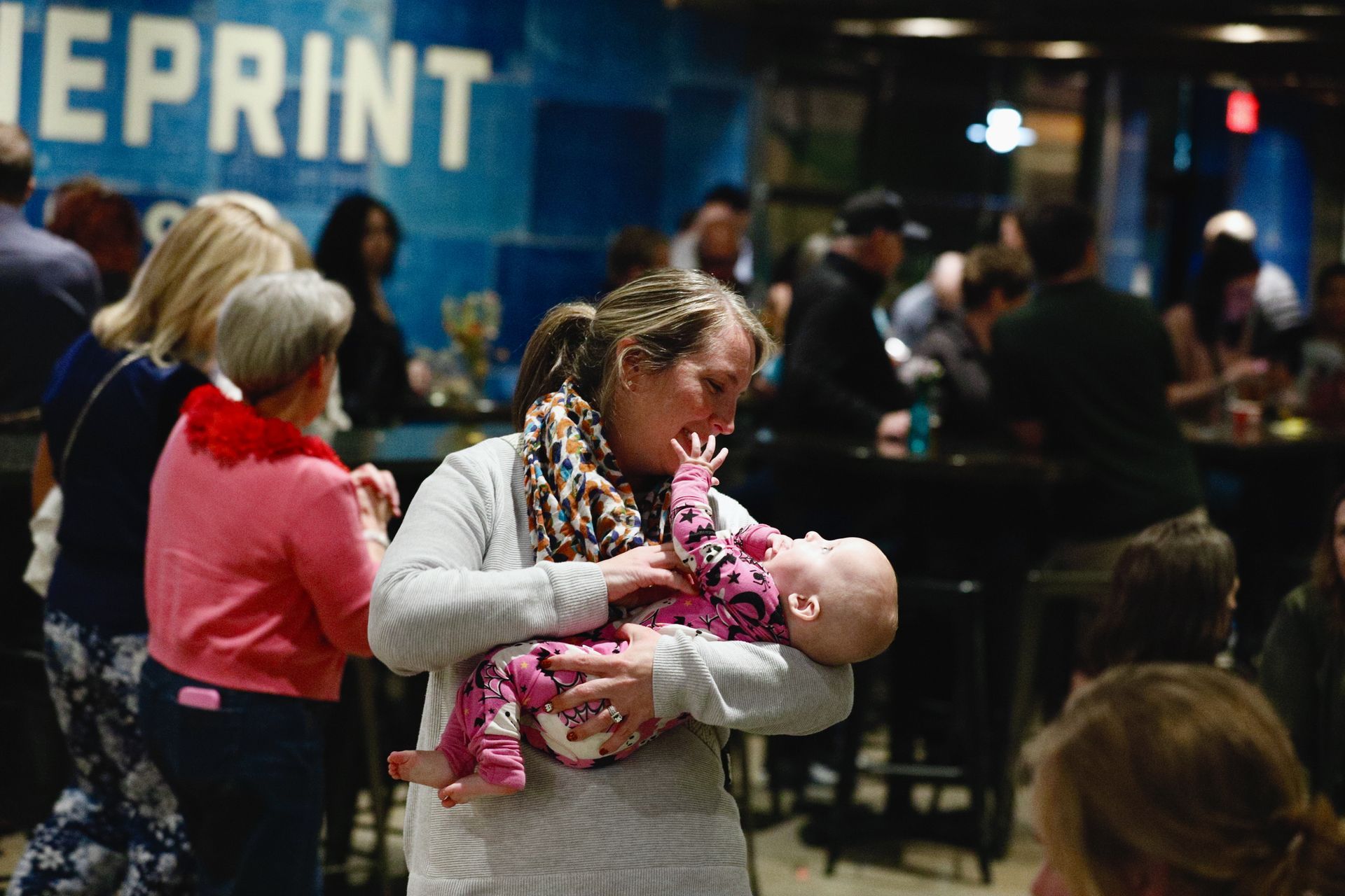 Woman holding baby in a crowded bar with blue wall. Baby is reaching up. Others are in the background.