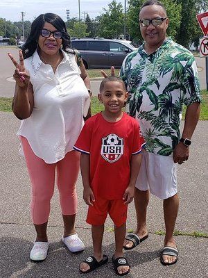 Family of three smiling outdoors: woman with peace sign, boy in red, man in tropical shirt.