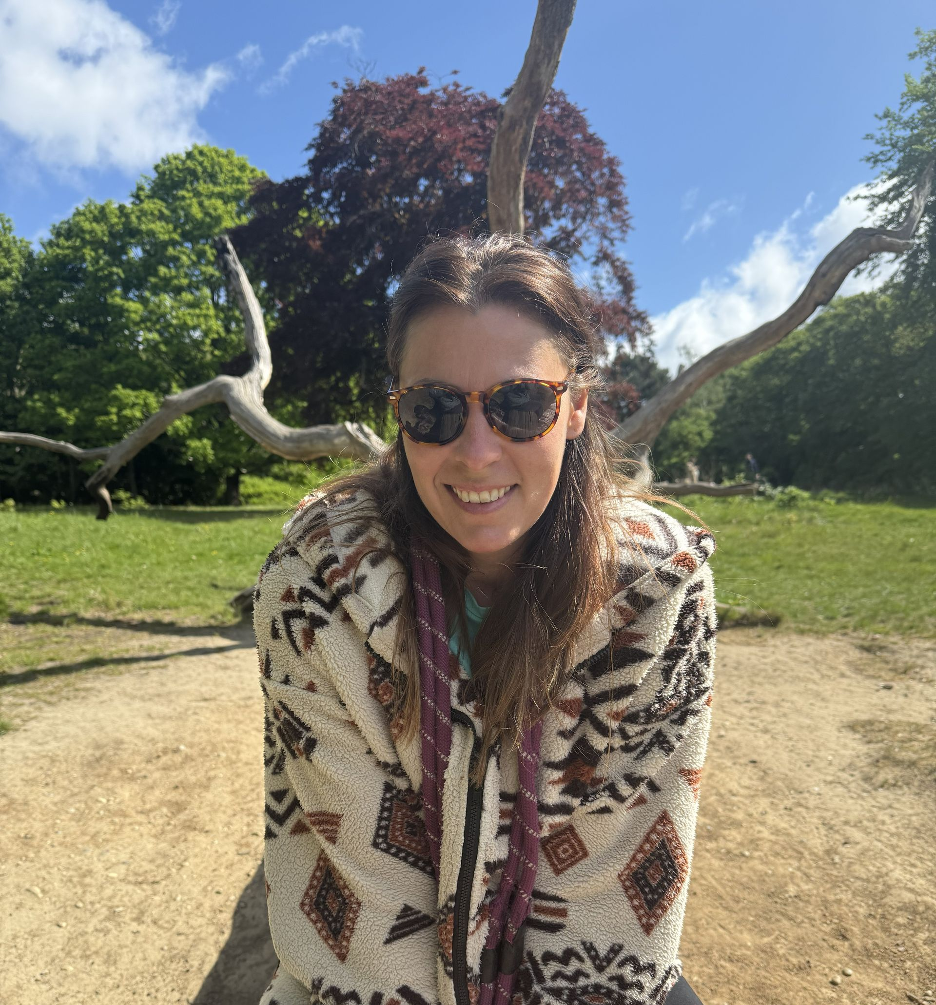 Woman in patterned coat and sunglasses smiles in a park setting, tree branches overhead.