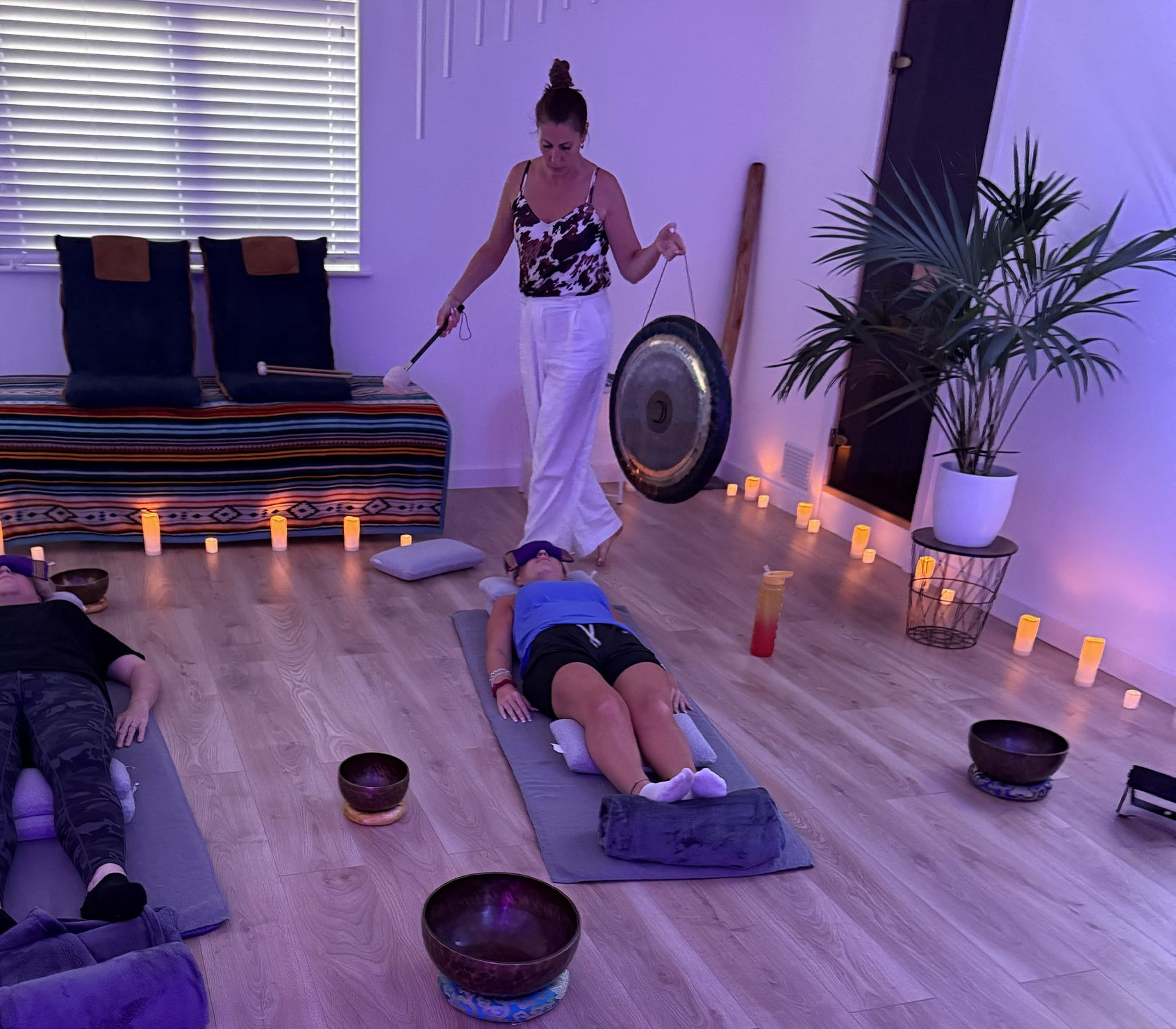 Woman playing gong in a dimly lit studio; people lying on mats with bowls around.