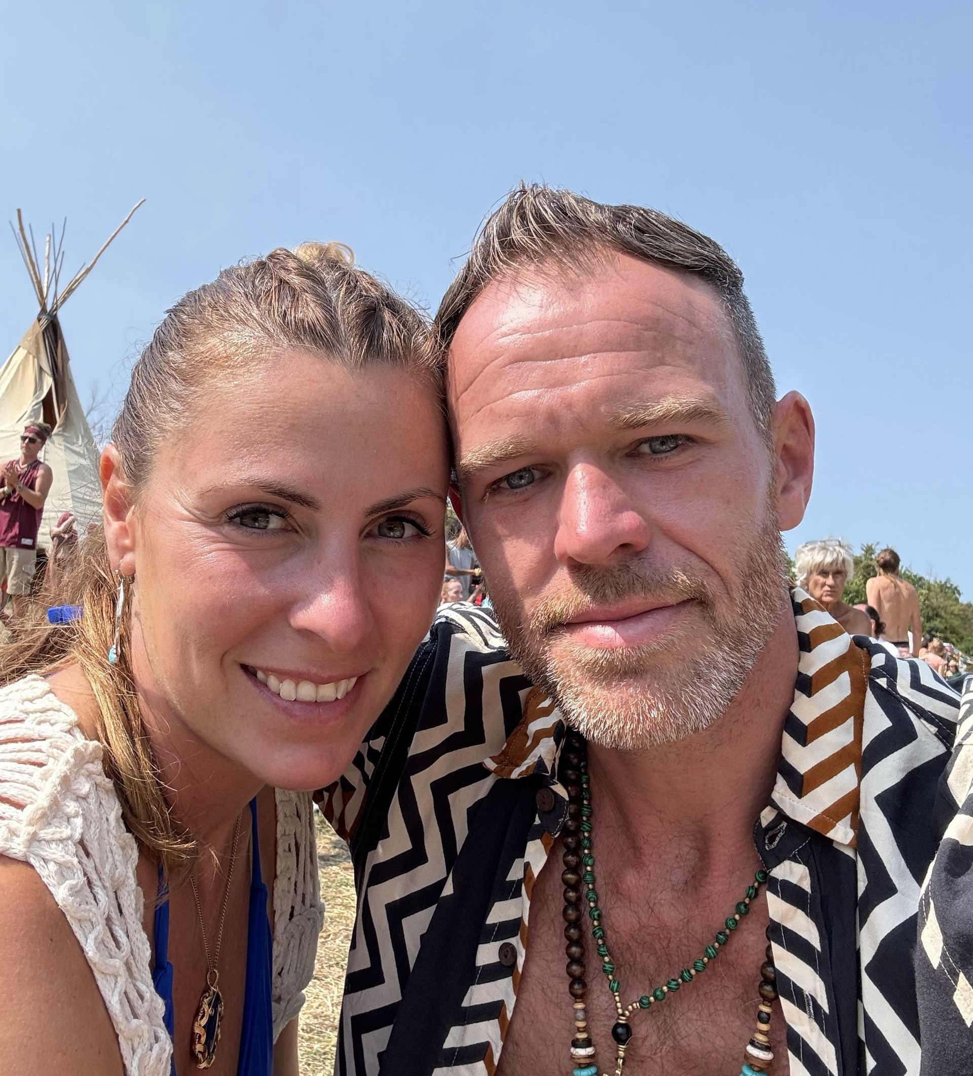 Couple smiling, posing close together outdoors. They are near a teepee with others visible in the background. Bright sunny day.