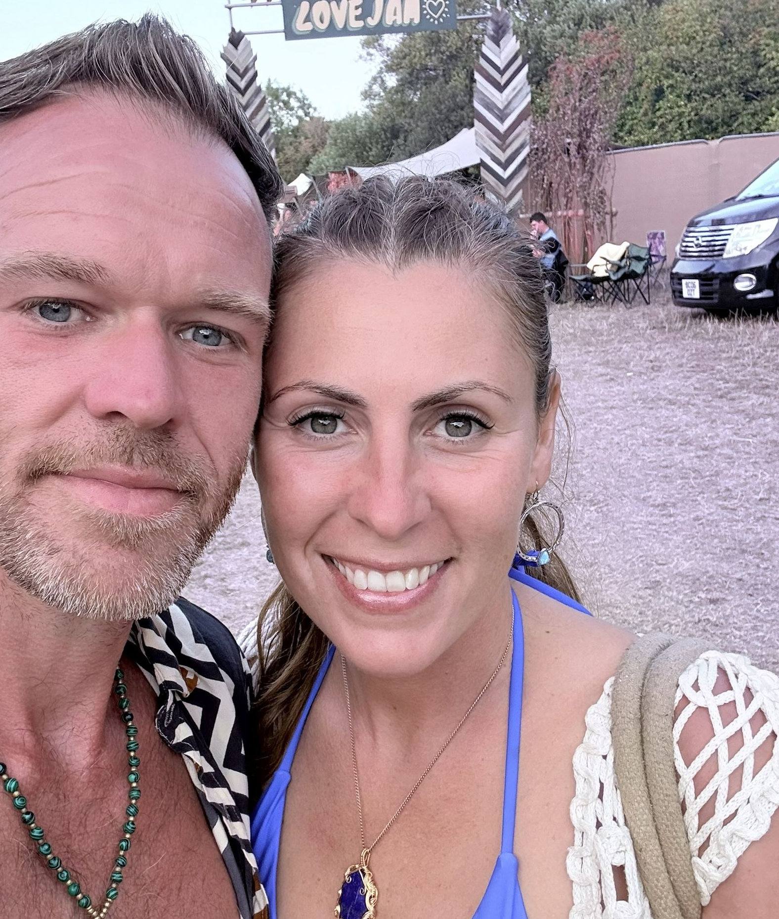 Couple smiling at a festival entrance. Man has graying stubble, woman in blue top. Love Jam sign visible.