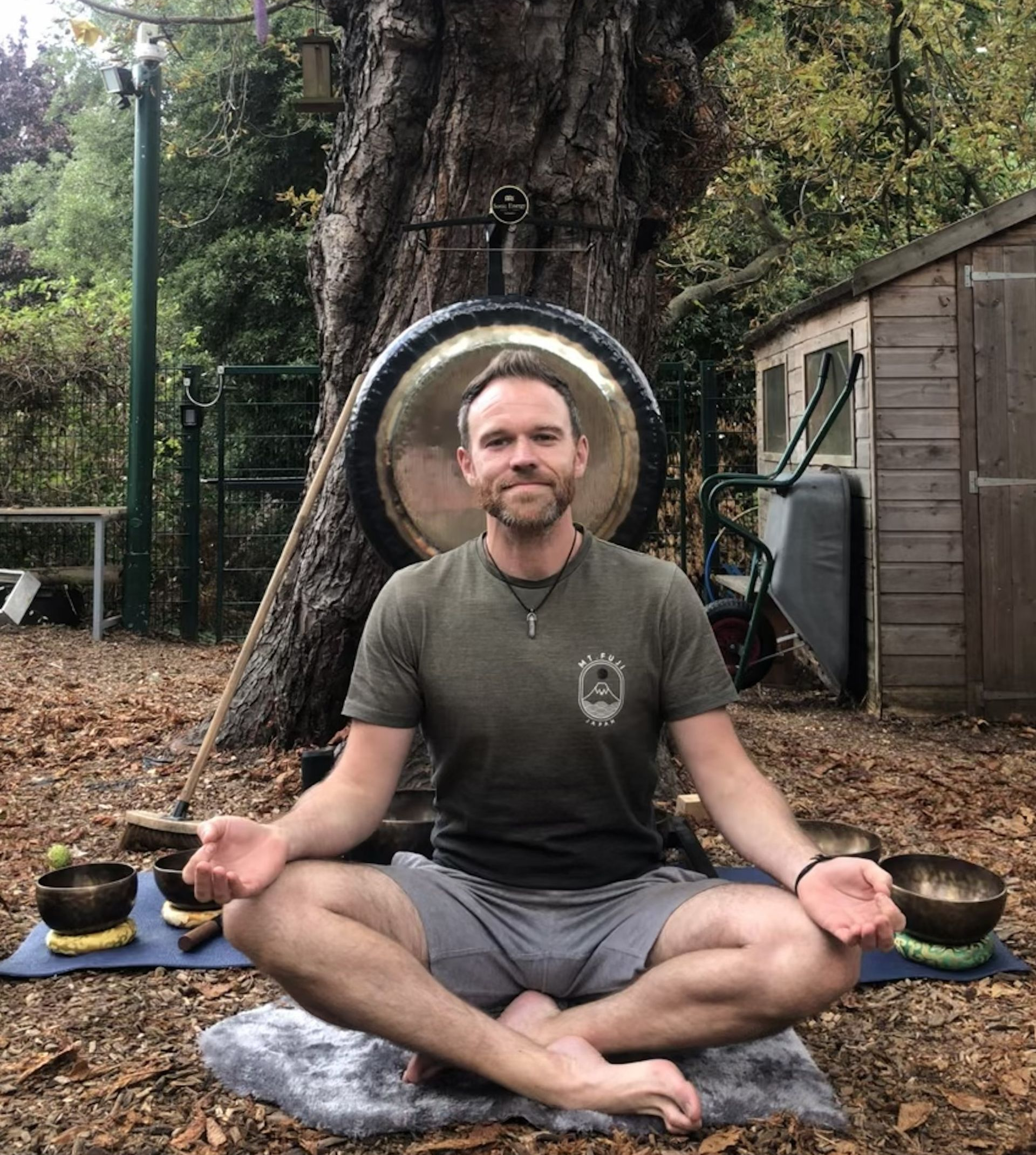 Man meditating outdoors with a gong, singing bowls, and shed in the background.