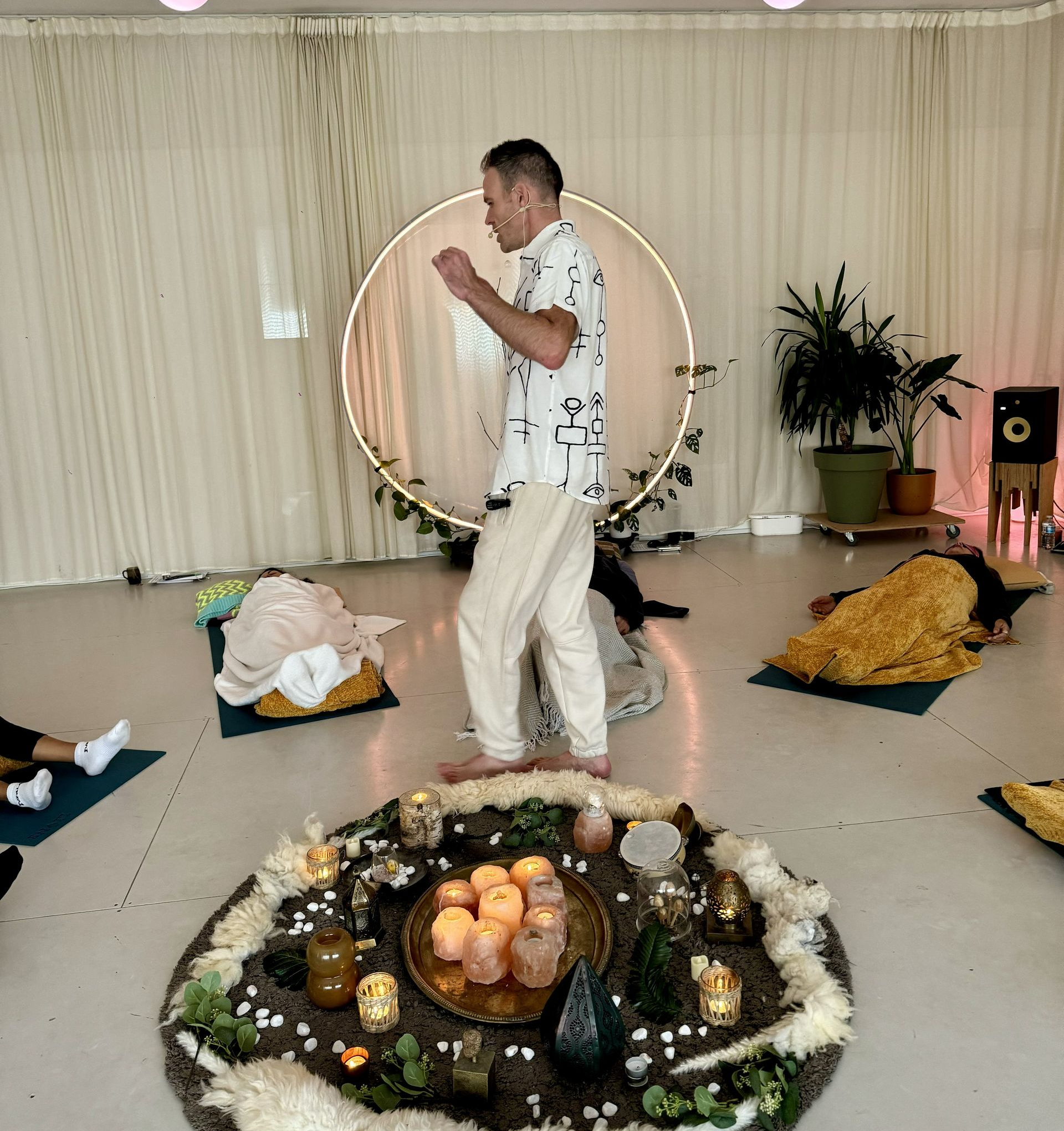 Man leads a group session; a circular lit frame and decorative circle on the floor, attendees lay on mats.