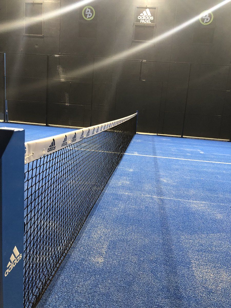 Campo da padel con superficie e rete blu. Logo Adidas visibile. Pareti nere sullo sfondo.