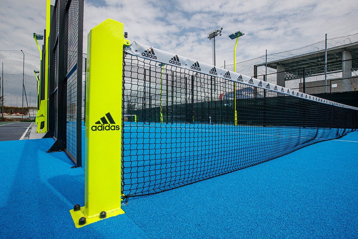 Campo da padel blu brillante con rete nera, palo giallo con logo Adidas. Stadio sullo sfondo.