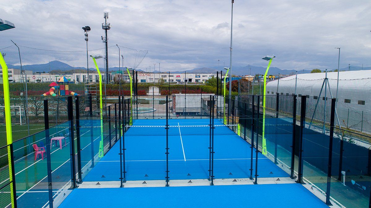 Campo da padel blu con pareti in vetro, recinzioni e luci, visto dall'alto sotto un cielo nuvoloso.