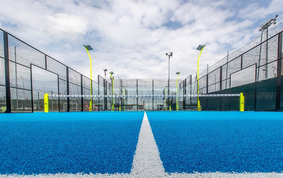 Campo da padel blu con linee bianche, pareti di vetro e una rete sotto un cielo nuvoloso.