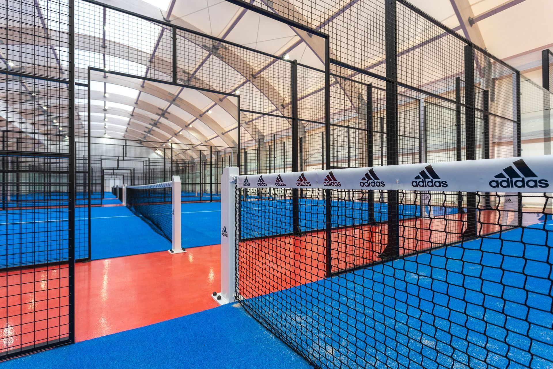 Campi da padel tennis al coperto con pavimentazione blu e rossa, rete nera e rete Adidas.