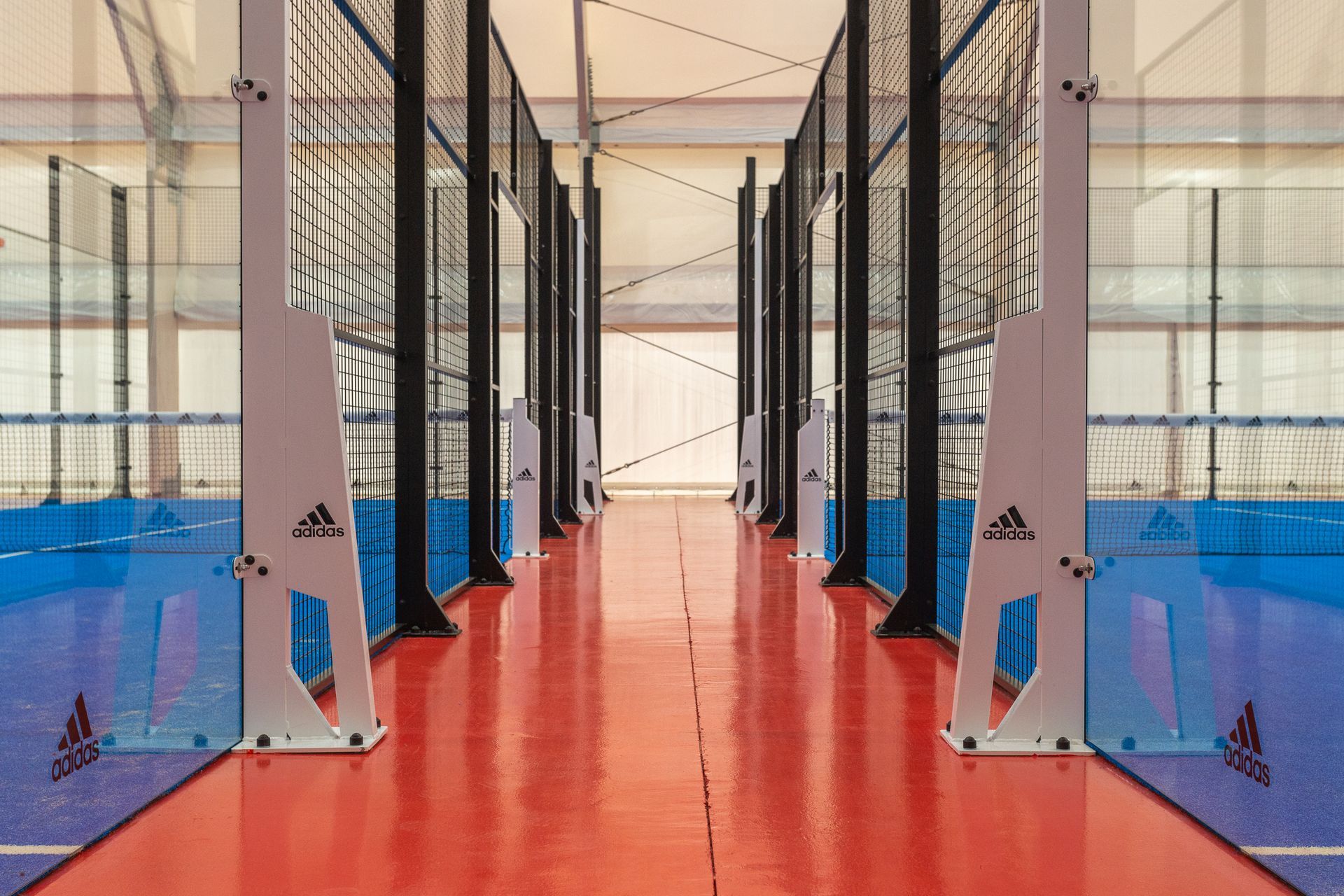 Campo da padel indoor con pavimentazione rossa e blu, pareti in vetro e marchio Adidas.