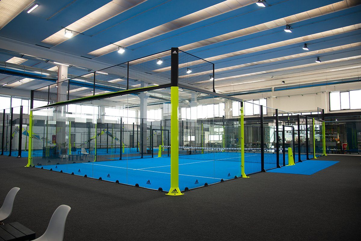 Campo da padel al coperto con pavimentazione blu, rete verde e nera e travi di supporto gialle.