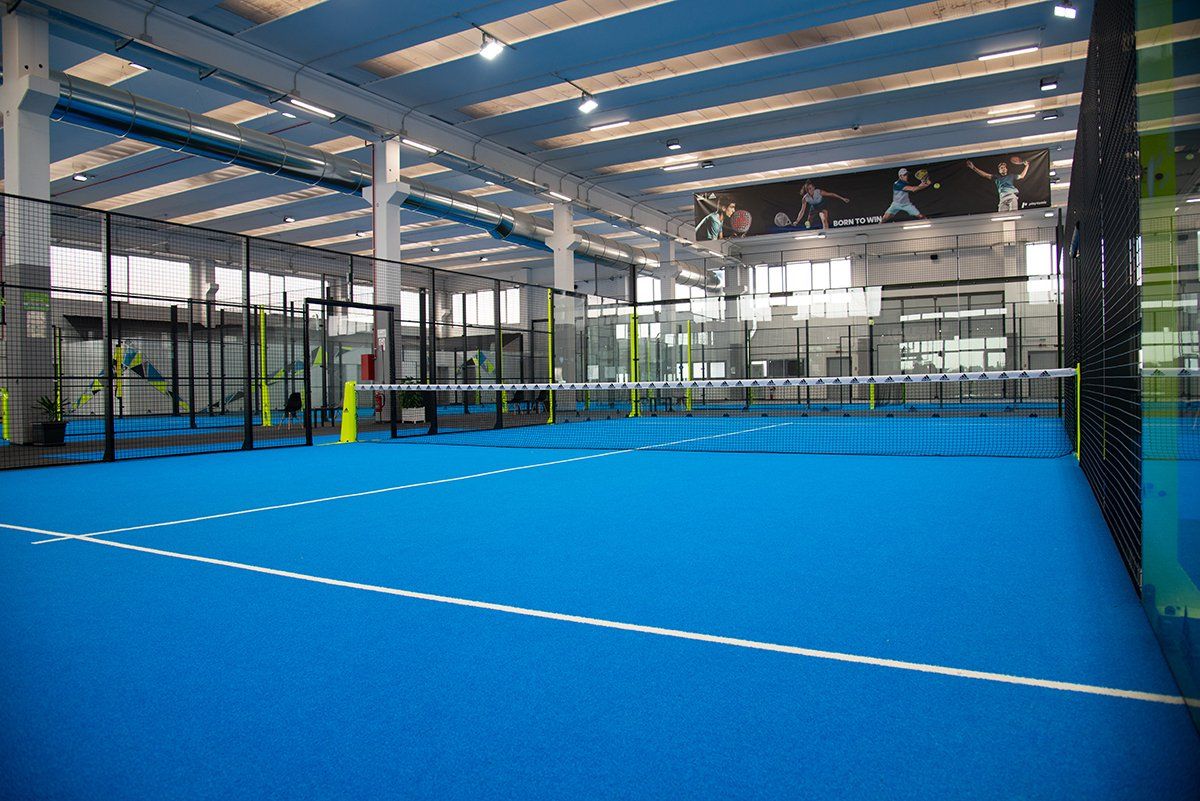 Campo da padel indoor con superficie blu e pareti in vetro, illuminato da luci intense.
