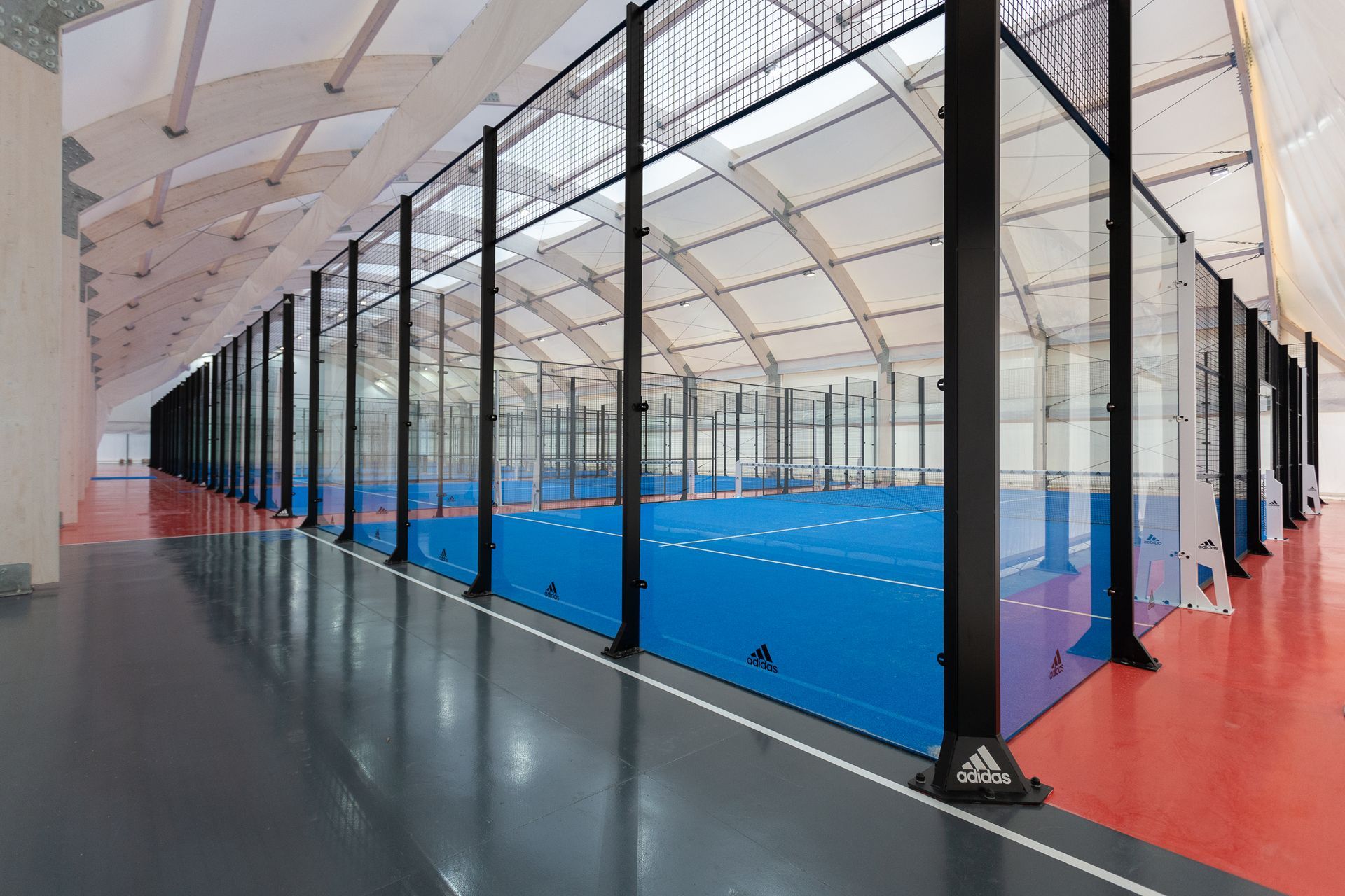 Campo da padel al coperto, superficie blu, pareti in vetro, travi di sostegno nere, pavimento grigio e rosso.