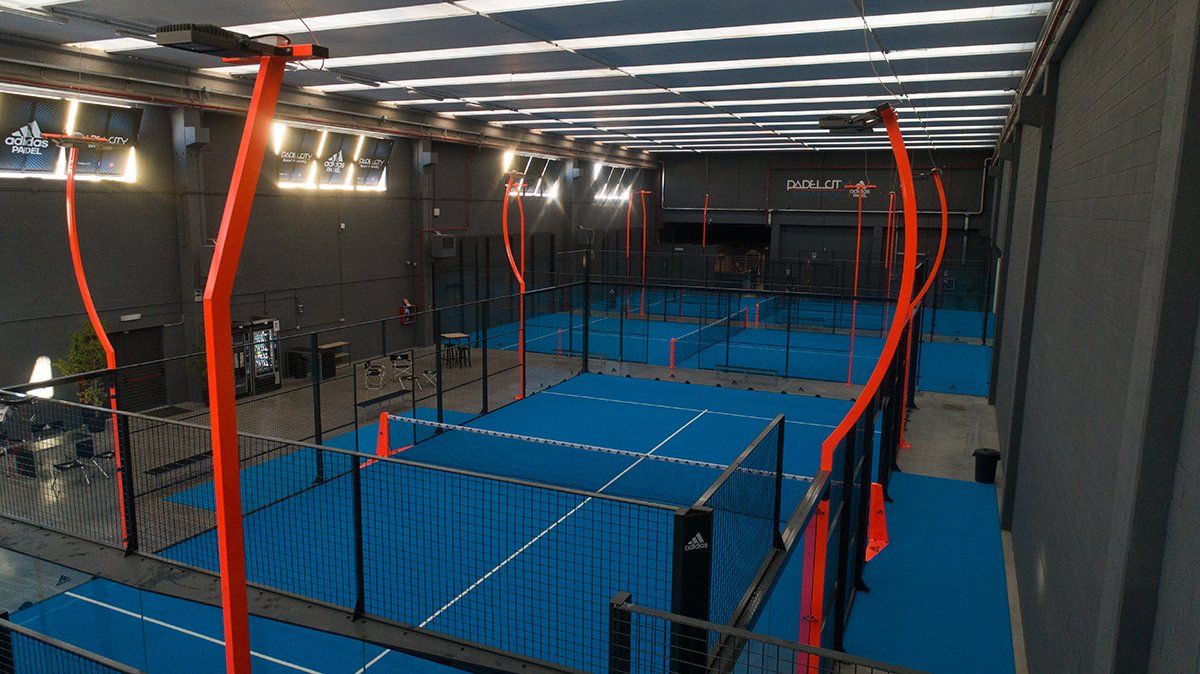 Campi da padel indoor con pavimentazione blu, telai neri e strutture di supporto arancioni.