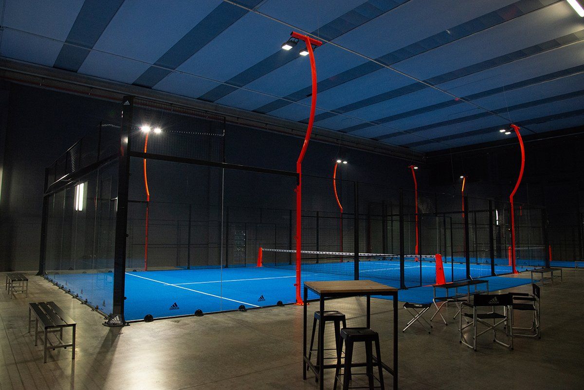 Campo da padel al coperto con pavimento blu, rete nera e pali rossi, illuminato da luci blu.