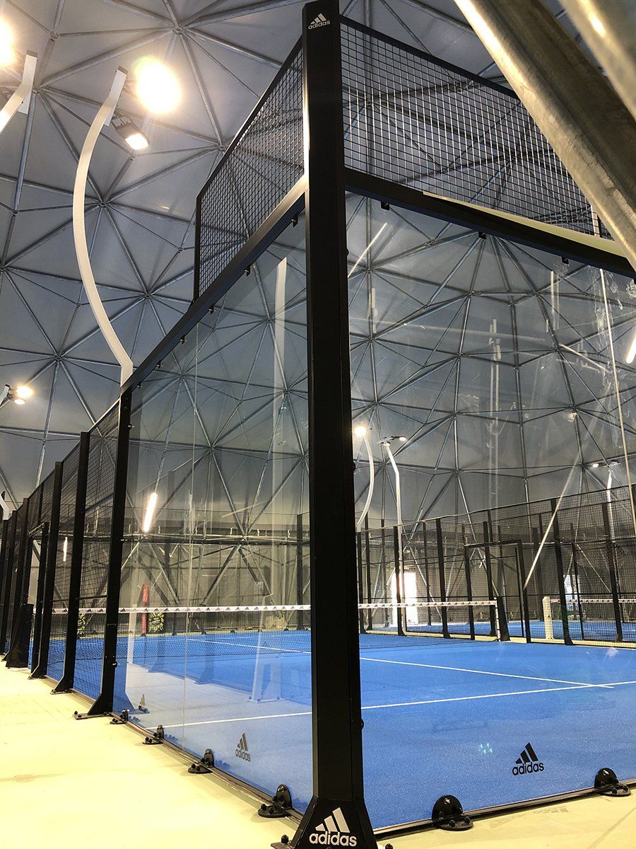 Campo da padel tennis racchiuso da pareti di vetro e strutture in metallo nero, all'interno di un edificio. 