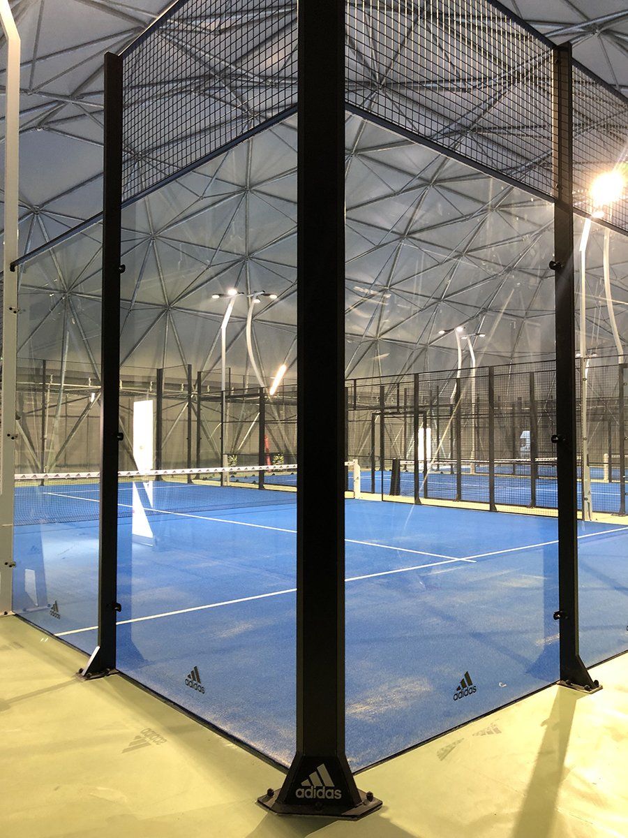 Campo da padel al coperto, superficie blu, struttura e rete nere, sotto un soffitto a cupola.